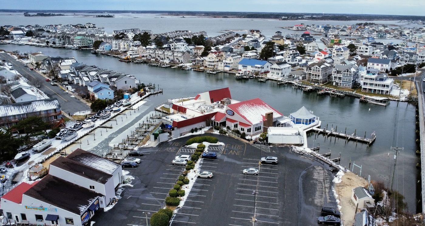 Drone photo of a waterfront restaurant with a red roof, its boat slips and large parking lot, with canal-front homes and the bay behind